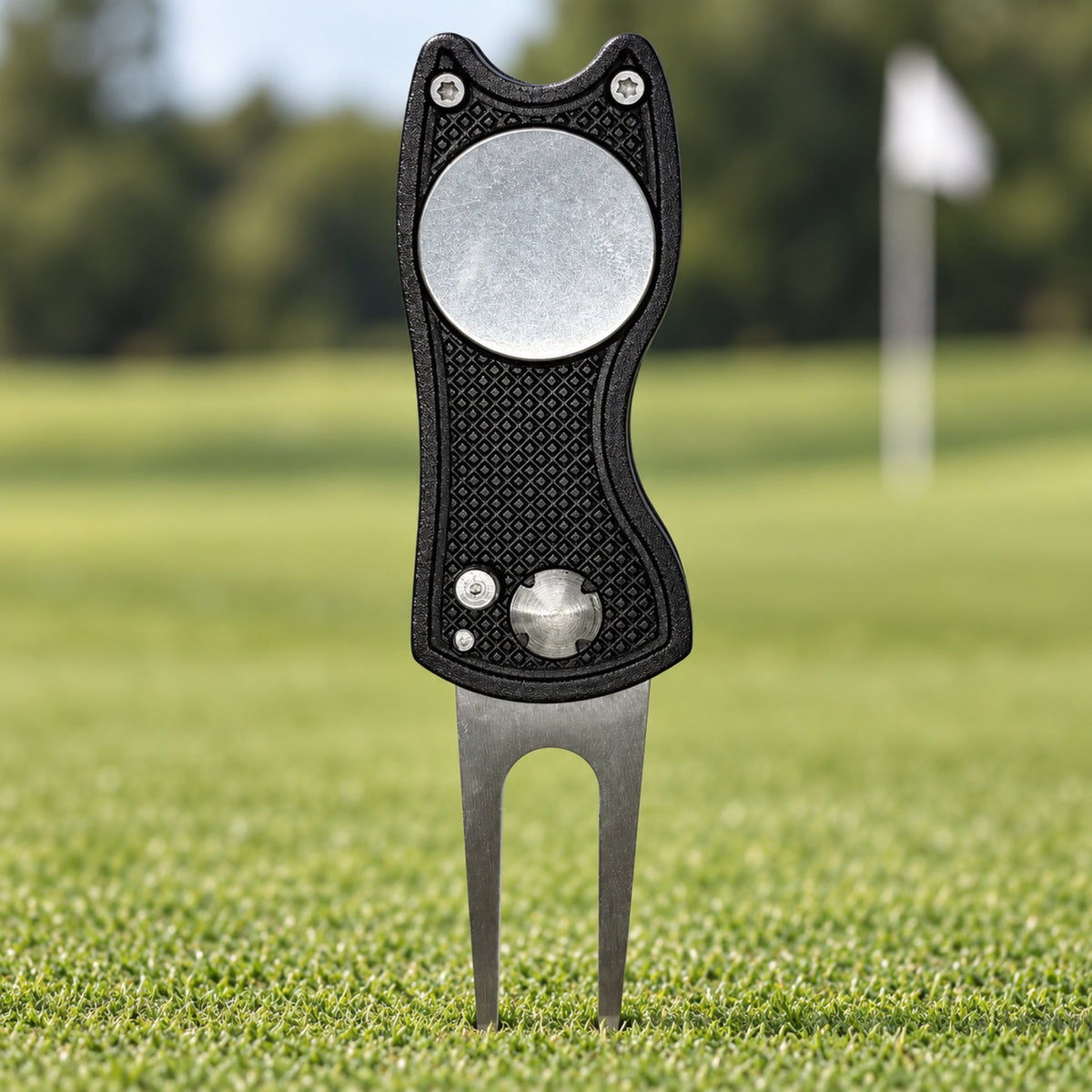 Black folding golf divot tool with a blank 25 mm ball marker, standing in a putting green