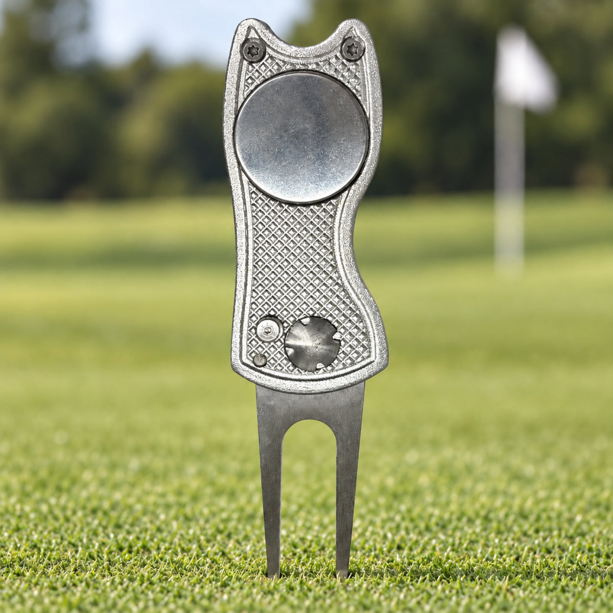 Chrome folding golf divot tool with a blank 25 mm ball marker, standing in a putting green